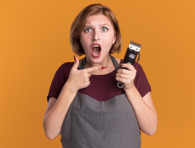 Young beautiful woman hairdresser in apron holding trimmer pointing with index finger at it being surprised and confused standing over orange wall