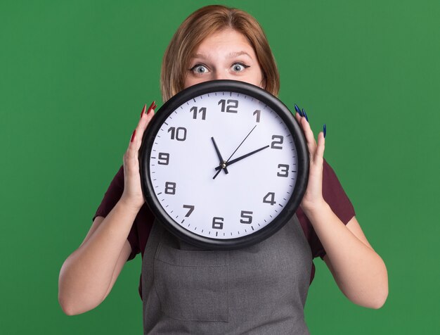 Young beautiful woman hairdresser in apron holding wall clock in front of her face peeking over standing over green wall