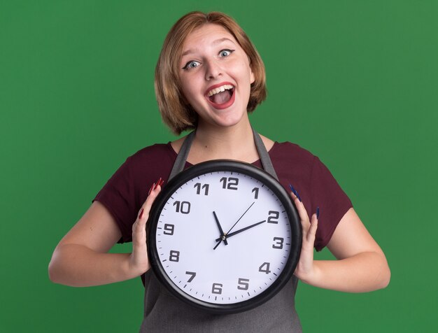 Young beautiful woman hairdresser in apron holding wall clock looking at front happy and cheerful smiling standing over green wall