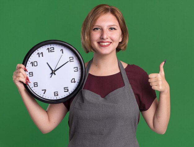 Young beautiful woman hairdresser in apron holding wall clock looking at front smiling cheerfully showing thumbs up standing over green wall