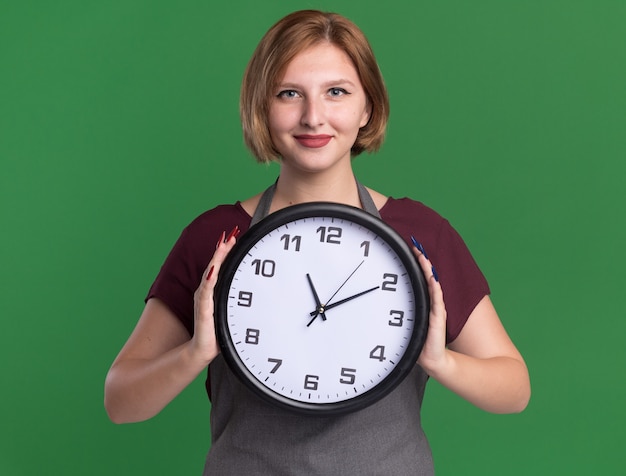 Giovane bella donna parrucchiere in grembiule tenendo l'orologio da parete guardando la parte anteriore con un sorriso fiducioso sul viso in piedi sopra la parete verde