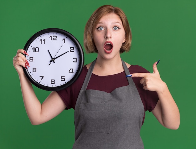 Young beautiful woman hairdresser in apron holding wall clock pointing with index finger at it amazed and surprised standing over green wall