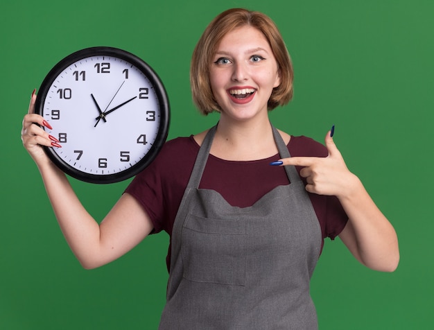 Young beautiful woman hairdresser in apron holding wall clock pointing with index finger at it smiling with happy face standing over green wall