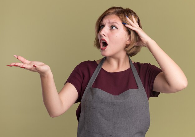 Young beautiful woman hairdresser in apron looking aside with arm out being confused and displeased standing over green wall