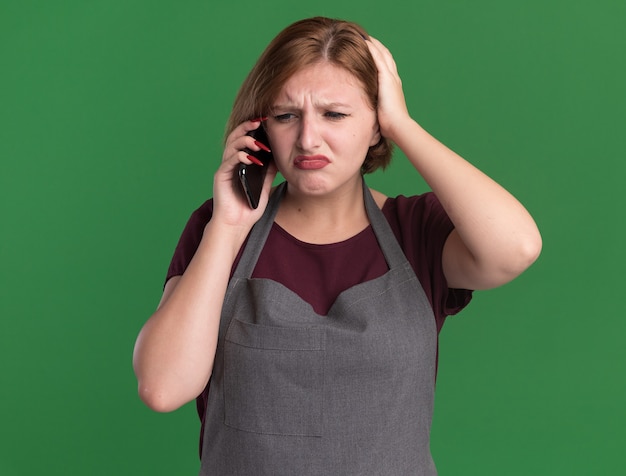 Young beautiful woman hairdresser in apron looking confused while talking on mobile phone standing over green wall