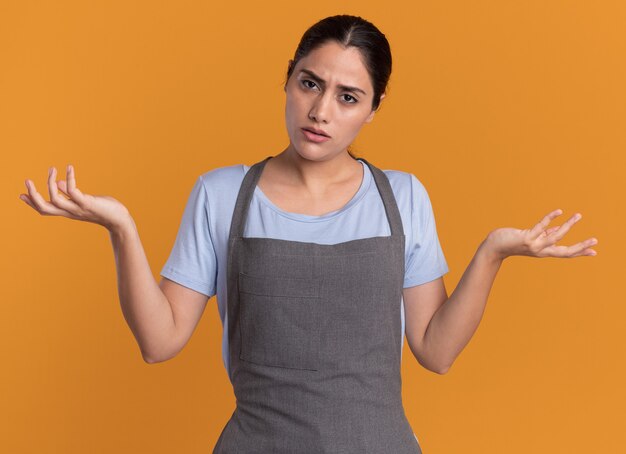 Young beautiful woman hairdresser in apron looking at front confused with arms raised having doubts standing over orange wall