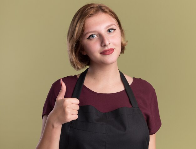 Young beautiful woman hairdresser in apron looking at front showing thumbs up smiling standing over green wall