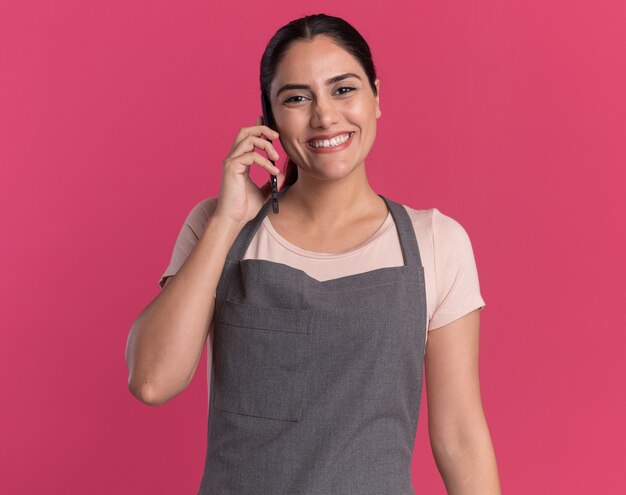 Young beautiful woman hairdresser in apron looking at front smiling with happy face while talking on mobile phone standing over pink wall