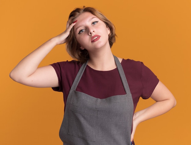 Young beautiful woman hairdresser in apron looking at front tired and bored with hand on her head standing over orange wall