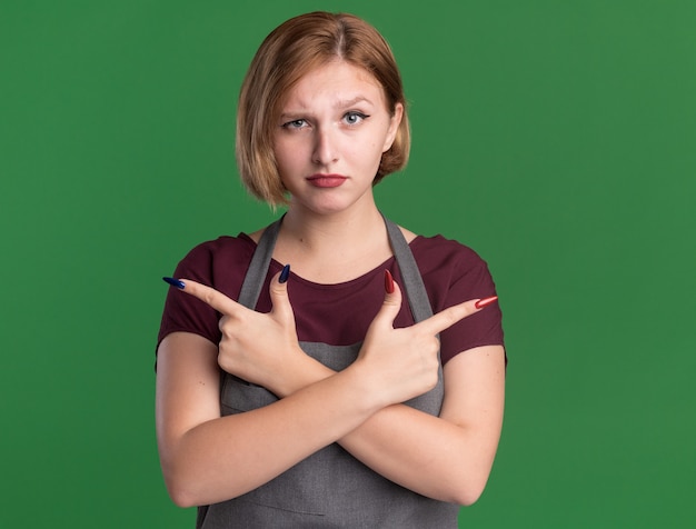 Parrucchiere di giovane bella donna in grembiule guardando davanti con faccia seria incrociando le mani che puntano con il dito indice ai lati in piedi sopra la parete verde