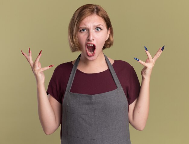 Young beautiful woman hairdresser in apron shouting with aggressive expression with arms raised standing over green wall