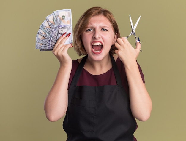 Young beautiful woman hairdresser in apron showing cash holding scissors shouting excited and confused standing over green wall