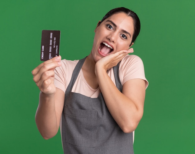 Young beautiful woman hairdresser in apron showing credit card happy and positive smiling standing over green wall