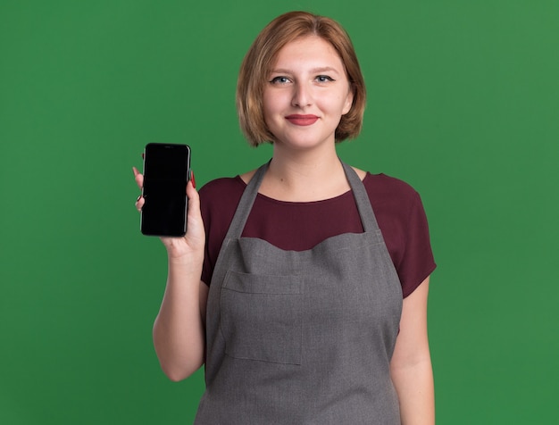 緑の壁の上に立って自信を持って笑顔のスマートフォンを示すエプロンの若い美しい女性の美容師