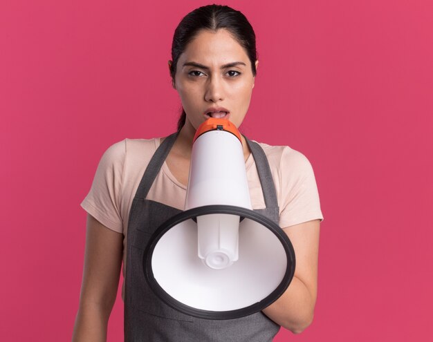 Young beautiful woman hairdresser in apron speaking to megaphone looking at front with serious face standing over pink wall