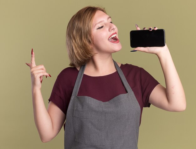 Молодая красивая женщина-парикмахер в фартуке с помощью смартфона в качестве микрофона поет счастливым и позитивным стоя над зеленой стеной