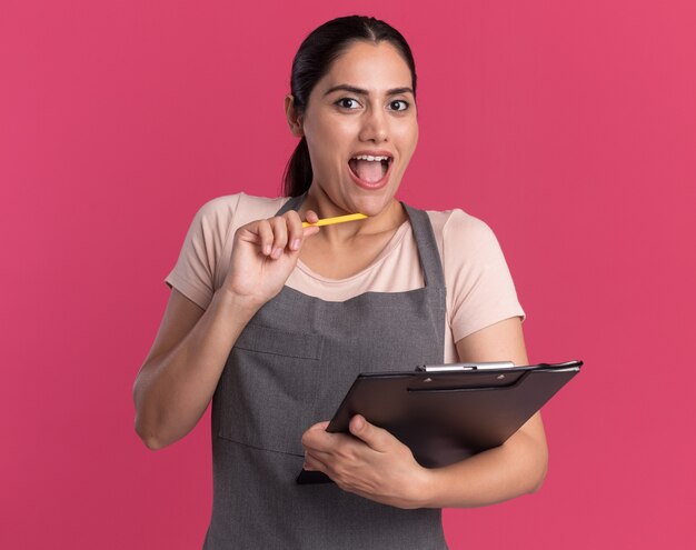 Young beautiful woman hairdresser in apron with pencil holding clipboard looking at front amazed and surprised standing over pink wall