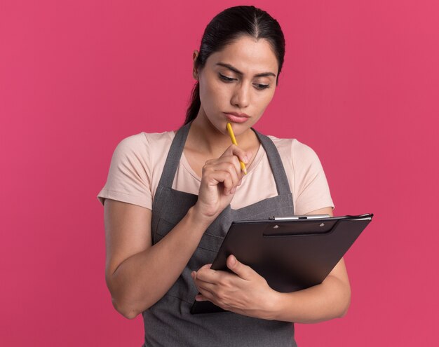 Young beautiful woman hairdresser in apron with pencil holding clipboard looking at it with serious face standing over pink wall