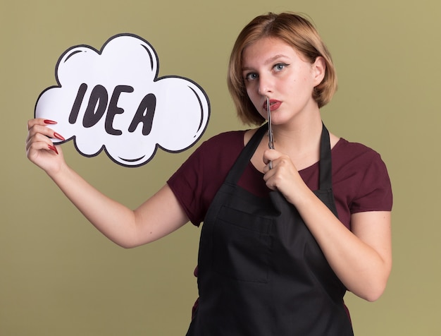 Young beautiful woman hairdresser in apron with scissors showing speech bubble sign with word idea looking at front with confident expression on smart face standing over green wall