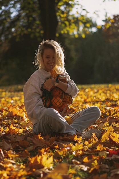秋の公園で帽子をかぶった若い美しい女性、オレンジのストリングバッグ、女性は紅葉を投げます。秋の気分、明るい自然の色。