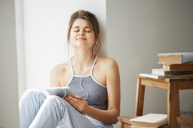 Young beautiful woman in headphones smiling with closed eyes holding tablet listening to streaming music sitting on floor over white wall.