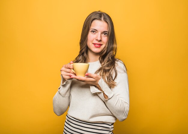 Young beautiful woman holding cup of coffee cup on hand standing against yellow backdrop