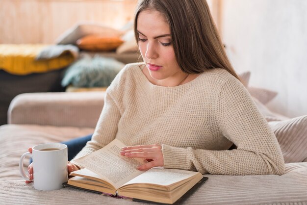 Young beautiful woman holding cup of coffee cup reading book