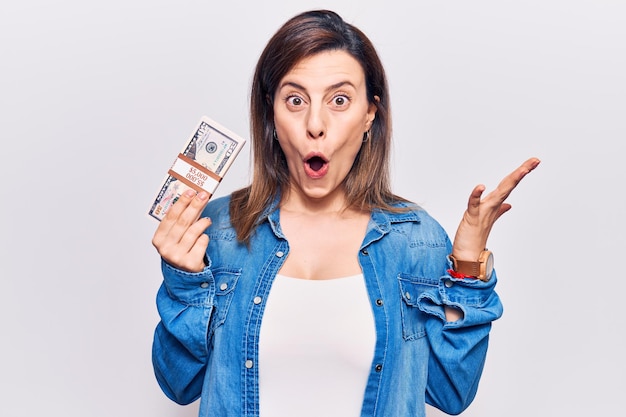 Young beautiful woman holding dollars scared and amazed with open mouth for surprise, disbelief face