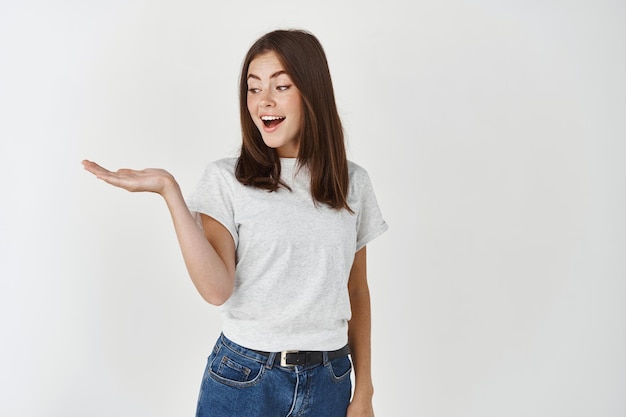 Young beautiful woman holding object on palm, extend hand on white wall and looking at product