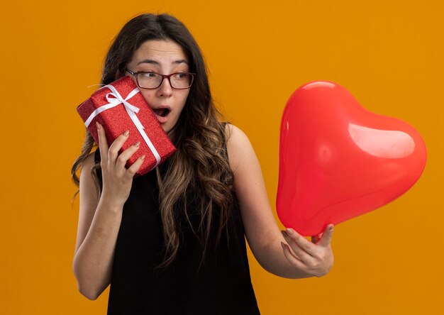 Young beautiful woman holding red balloon in heart shape and gift looking surprised and happy smiling cheerfully celebrating valentine's day