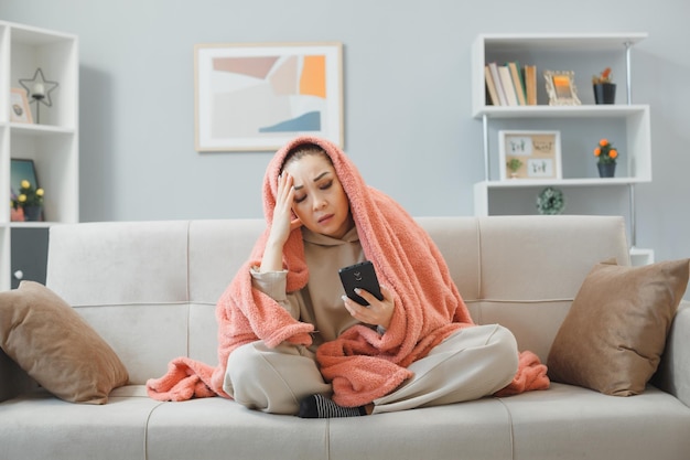 Молодая красивая женщина в домашней одежде сидит на диване под одеялом в домашнем интерьере с помощью смартфона и выглядит нездоровой с головной болью