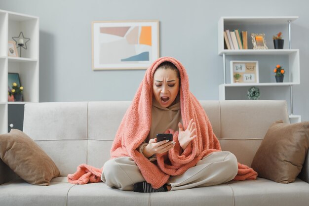 Молодая красивая женщина в домашней одежде сидит на диване под одеялом и читает сообщение с помощью смартфона, выглядит злой и разочарованной