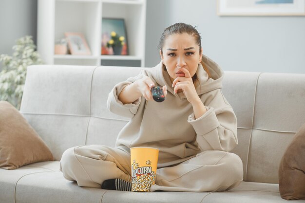 young beautiful woman in home clothes sitting on a couch at home interior with bucket of popcorn holding remote watching television being confused and displeased