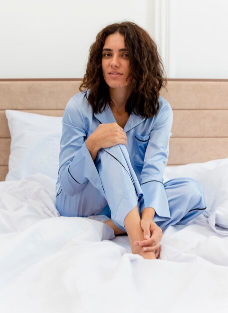 Бесплатное фото Молодая красивая женщина в синей пижаме сидит на кровати, глядя в камеру с улыбкой на лице в интерьере спальни на светлом фоне