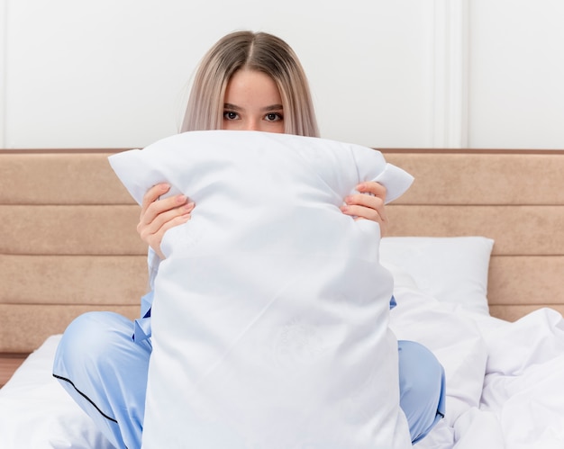 Бесплатное фото Молодая красивая женщина в синей пижаме сидит на кровати с подушкой, пряча лицо, выглядывает в интерьере спальни