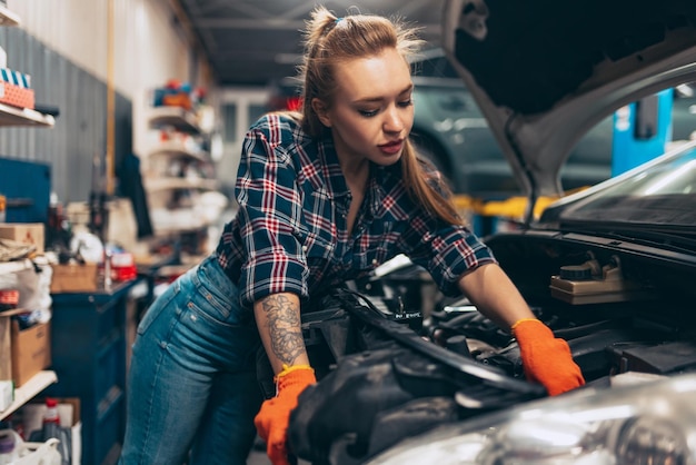 Бесплатное фото Молодая красивая женщина в клетчатой рубашке, работающая автомехаником, чинит машину на автосервисе
