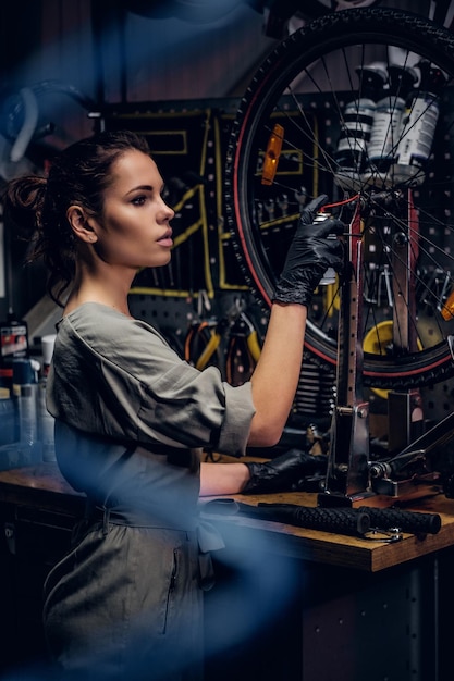 Free photo young beautiful woman is repairing bicycle at busy workshop between pneumatic wires.