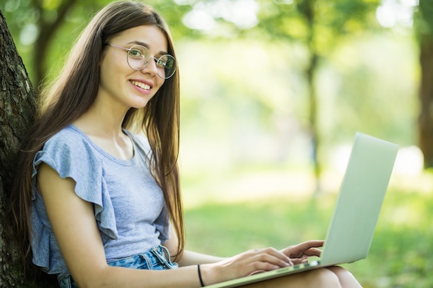 Young beautiful woman is sitting on green grass under the tree in the garden on summer day and working on her laptop
