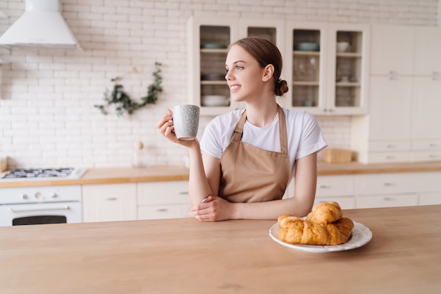 コーヒーとクロワッサンを持ったエプロンの台所で若い美しい女性が朝を楽しんでいる