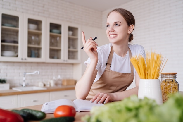 Young beautiful woman in the kitchen in an apron writes down her favorite recipes next to fresh vegetables