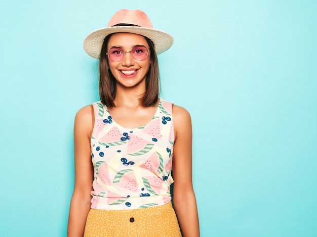 Young beautiful woman looking at camera in hat. Trendy girl in casual summer white T-shirt and yellow skirt in round sunglasses. Positive female shows facial emotions. Funny model isolated on blue