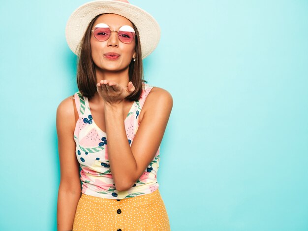 Young beautiful woman looking at camera in hat. Trendy girl in casual summer white T-shirt and yellow skirt in round sunglasses. Positive female shows facial emotions. Gives air kiss