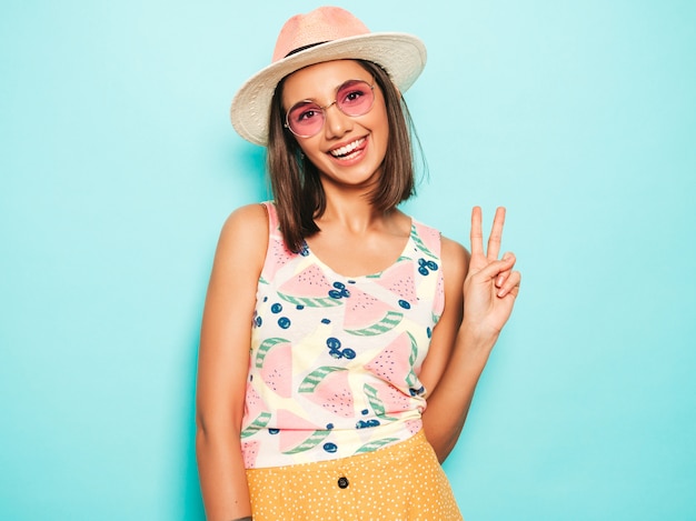 Free photo young beautiful woman looking at camera in hat. trendy girl in casual summer white t-shirt and yellow skirt in round sunglasses. positive female shows facial emotions. shows peace sign