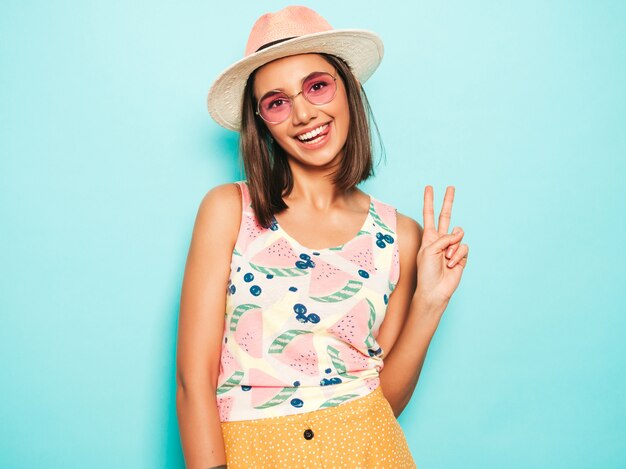 Young beautiful woman looking at camera in hat. Trendy girl in casual summer white T-shirt and yellow skirt in round sunglasses. Positive female shows facial emotions. Shows peace sign