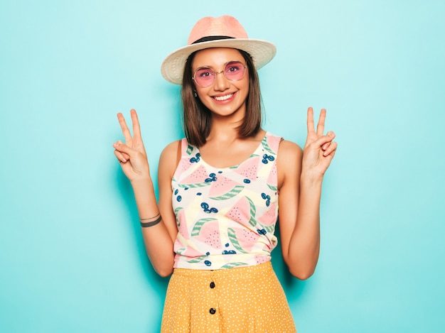 Young beautiful woman looking at camera in hat. Trendy girl in casual summer white T-shirt and yellow skirt in round sunglasses. Positive female shows facial emotions. Shows peace sign