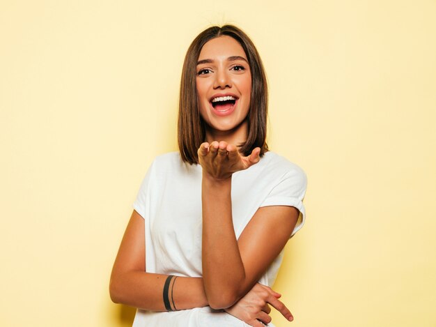 Young beautiful woman looking at camera. Trendy girl in casual summer white T-shirt and jeans shorts. Positive female shows facial emotions. Funny model isolated on yellow gives air kiss