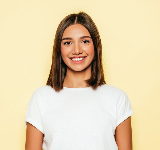 Young beautiful woman looking at camera. Trendy girl in casual summer white T-shirt and jeans shorts. Positive female shows facial emotions. Funny model isolated on yellow
