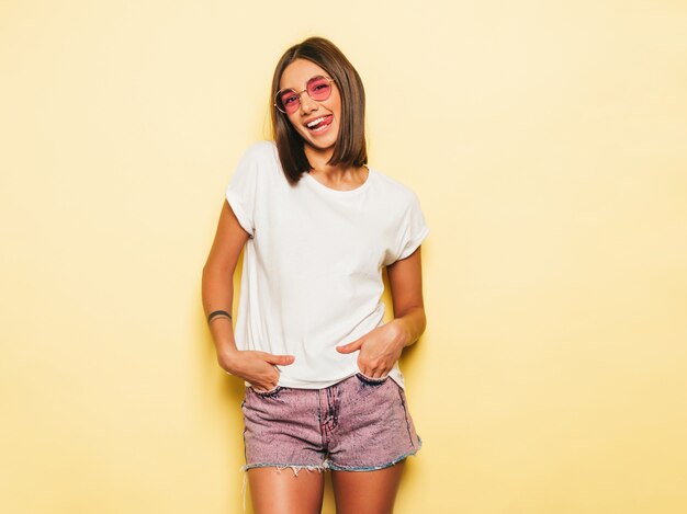 Young beautiful woman looking at camera. Trendy girl in casual summer white T-shirt and jeans shorts in round sunglasses. Positive female shows facial emotions. Model isolated on yellow shows tongue