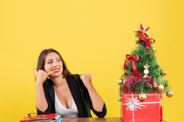 Молодая красивая женщина делает жест «позвони мне», сидя за столом возле украшенной елки в офисе на желтом
