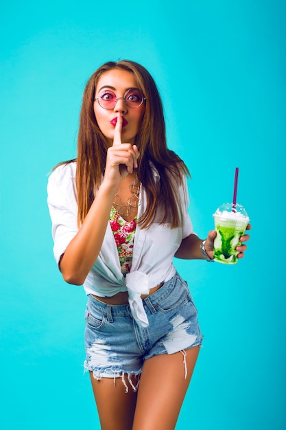 Young beautiful woman in mini denim shorts drinking tasty smoothie, vintage outfit, make up sunglasses